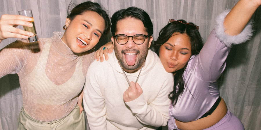 Three friends posing closely together at a party, smiling and laughing while holding drinks in front of a light curtain backdrop.