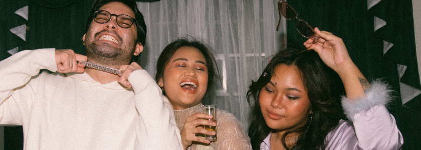 Three friends celebrating at a party indoors, smiling and dancing while holding drinks in front of a curtained window backdrop.
