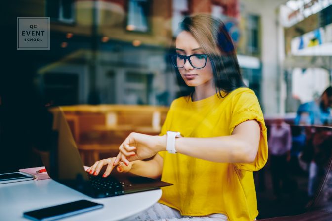 Young businesswoman in eyeglasses looking at display of wristwatch and managing time while checking email on laptop.Freelancer with smartwatch on hand working on article at netbook before deadline