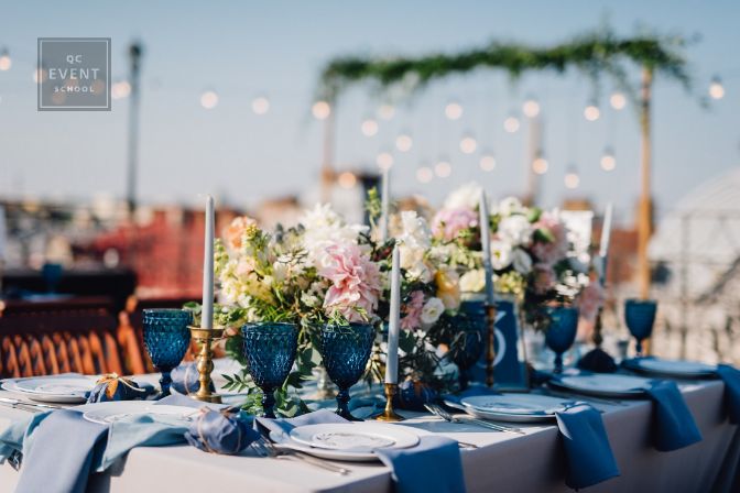 Long dinner tables covered with grey and blue cloth, served with porcelain and blue glasses and rich decorated with flowers stand on the roof of a house. Bokeh lights on background
