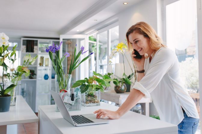 Female florist standing at her flower shop counter using mobile phone and laptop to take orders for her store. Woman flower shop owner taking online orders.