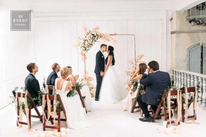 Stylish wedding ceremony decorated with flowers and green branches. Couple bride and groom near the arch hold hands and kiss, guests are sitting on chairs
