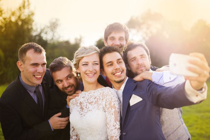 Outdoor portrait of beautiful young bride with groom and his friends taking selfie. Virtual wedding article.