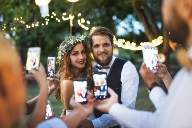 Guests with smartphones taking photo of bride and groom at wedding reception outside. Virtual weddings article.