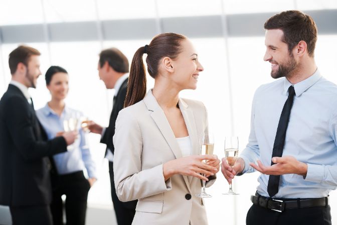 Celebrating their common success. Two cheerful business people drinking champagne and talking while other people communicating in the background. Corporate event planning article.