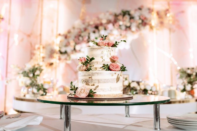 The wedding cake. White, two-tiered, decorated with flowers and gold. On a delicate pink background. Side view, top. Wedding planner myths article.