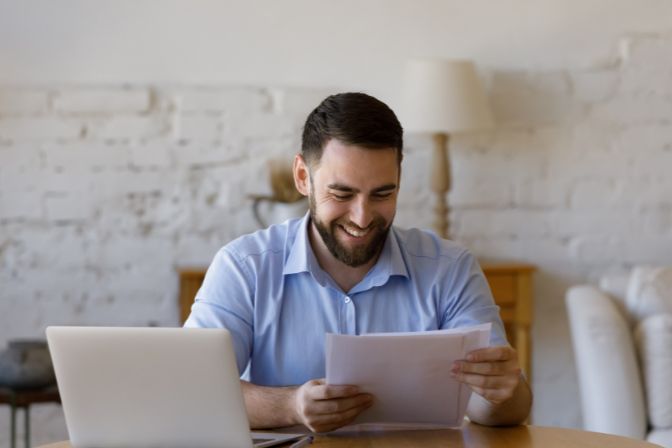 Handsome business man sit at table with laptop holding papers, read received contract, agreement, legal documents smile feel satisfied, got good news. Loan repayment, career advance, success concept. Contract article.