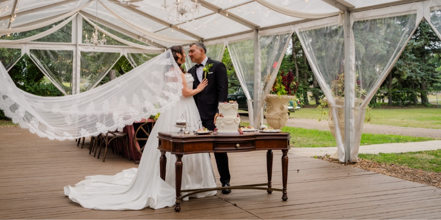 A bride and groom looking at each other inside a clear event tent with white draped fabric and chandeliers, standing behind a wooden vintage table holding a white wedding cake, as the bride's long lace-edged veil flows in the breeze.