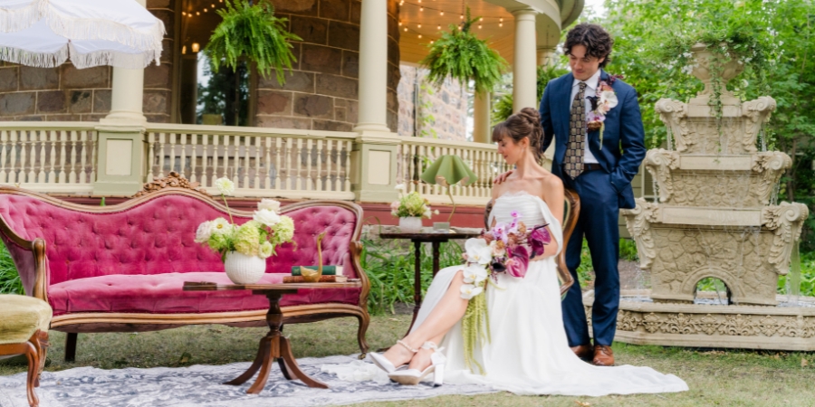 A bride and groom posing in a vintage outdoor wedding lounge setting featuring a tufted magenta velvet sofa, a patterned rug on the grass, and a tiered stone fountain.