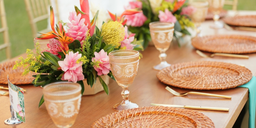 A beautifully set wooden event table featuring vibrant tropical floral centerpieces, woven rattan charger plates, gold flatware, and ornate amber glass goblets.